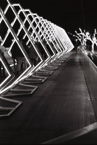 Black and white photograph of a modern geometric walkway at night, featuring a series of illuminated diamond-shaped frames on the left side and blurred figures walking along the path.